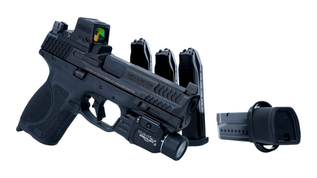Black semi-automatic handgun with red-dot sight, tactical flashlight attached, three curved magazines, and a detachable holster/attachment against a white background